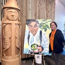 A women stands next to the painting of her father and a carving the represents her brother