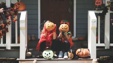 Little children in Halloween costumes sitting on steps holding pumpkins in front of their faces