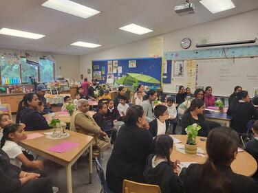 Classroom filled with students and family members sitting in chairs listening to a presentation.