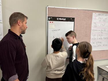Three students working on math program hung on wall while teachers observes.