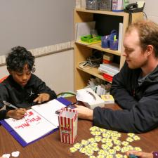 Male Educational Assistant sits with male elementary student playing education game at a table