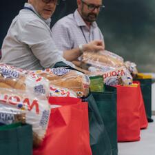 Two male staff standing at tables with food hampers that are being handed out to families