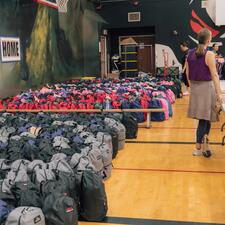 Rows and rows of filled backpacks lined up on the gymnasium floor, up against the wall as a staff member stands and waits for students to pick one up.